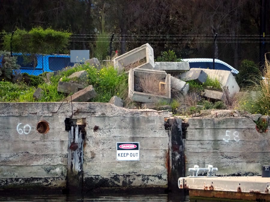 A concreted side of a river with weeds growing amongst a pile of rubble.
