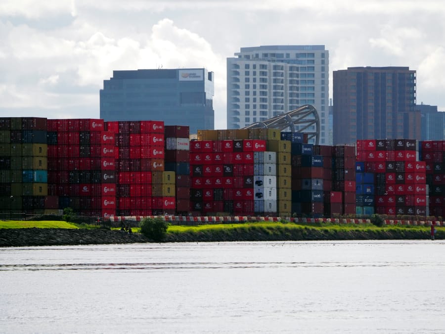 River in foreground, stacked shipping containers along the shore and three high-rise buildings in the background.